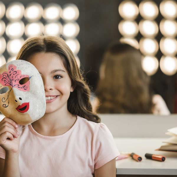 nina-sonriente-sentada-backstage-sosteniendo-mascara-delante-su-cara